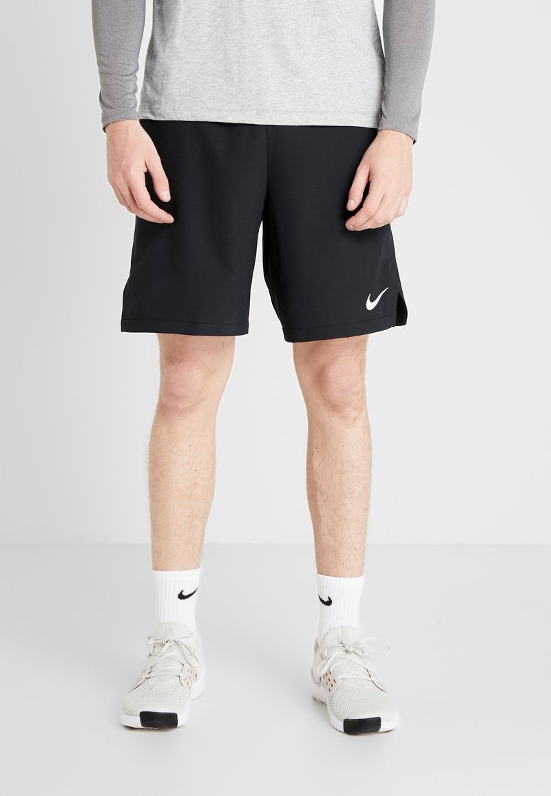 Homme portant un short Nike noir, des chaussettes blanches Nike et des baskets gris clair, debout contre un fond clair uni.