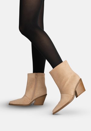 Piernas con medias negras que llevan botines beige de ante con tacones gruesos apilados y cremalleras laterales sobre un fondo blanco.