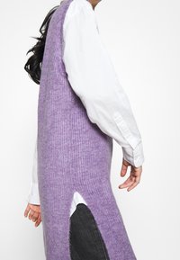 Lila gestricktes ärmelloses Kleid mit einer weichen Textur, das eine seitliche Schlitze hat und über einem weißen Hemd mit Knöpfen getragen wird.