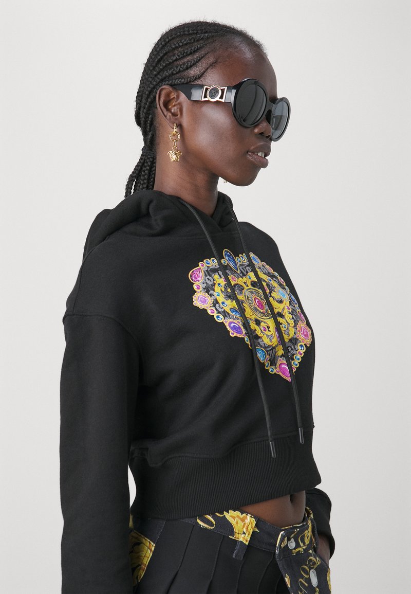 Haut de survêtement noir court avec un design graphique coloré, arborant des motifs ornés en jaune, rose et bleu. Associé à des lunettes de soleil rondes noires.