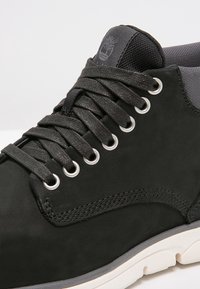 Svart syntetiskt läder sneakers med rund tå, grå textilkonton, silverfärgade öglor och svarta snören. Har en texturerad, upphöjd logotyp.