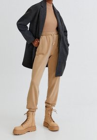 Long manteau gris à carreaux sur un haut côtelé beige, associé à un pantalon beige à taille élastique et des bottines cognac à lacets avec une semelle épaisse.