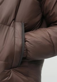 Brązowa kurtka puchowa z pikowanym wzorem, elastycznymi mankietami i boczną kieszenią. Wykonana z lekkiego, wodoodpornego materiału.