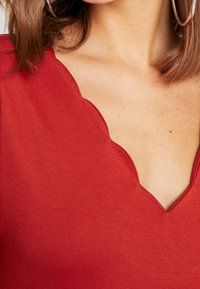 Detailní záběr na ženu v červeném topu s vroubkovaným výstřihem do V a velkými kruhovými náušnicemi, který ukazuje její krk a horní část hrudníku.