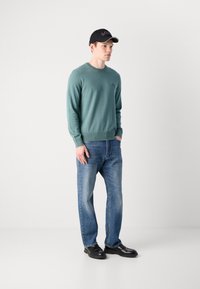 Young man wearing a teal sweater, blue jeans, black leather shoes, and a black cap, standing against a plain white background.