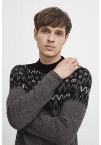 Grauer und schwarzer Strickpullover mit einem gemusterten Schulterbereich, der geometrische Designs und einen hohen Kragen aufweist. Der Stoff wirkt weich und strukturiert.