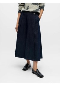 Jupe midi en denim bleu foncé avec une silhouette structurée, poche avant, détails de couture sur le côté et fermeture à bouton métallique. Associée à des mocassins noirs.