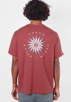 Personne portant un t-shirt couleur rouille avec un soleil argenté circulaire et le texte "OXBOW" dans le dos sur un fond uni.