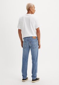 Hombre con el pelo corto decolorado que lleva una camiseta blanca, jeans azul claro y zapatillas negras, de pie de espaldas sobre un fondo blanco.