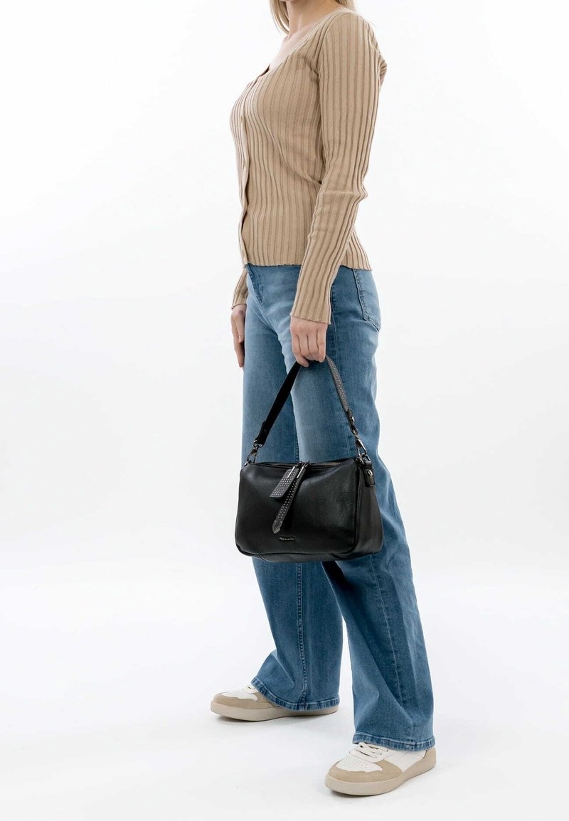 Personne en pull côtelé beige et jean bleu tenant un sac à main en cuir noir, debout devant un fond blanc uni.