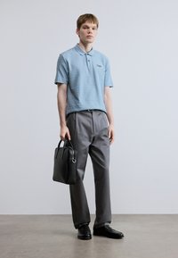 Φωτεινό μπλε πόλο μπλουζάκι με υφασμάτινες ρίγες, γκρι παντελόνι και μαύρα δερμάτινα παπούτσια. Κρατάει μια μαύρη τσάντα χειρός με διπλά χερούλια.