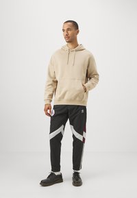 Sudadera beige con bolsillo canguro y cordón; pantalones de chándal negros con rayas diagonales blancas y burdeos; zapatos negros.