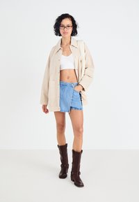 Beige lærredjakke, hvid crop top, denim omslagskørter og brune knæhøje støvler med spænde detaljer. Model står foran en neutral baggrund.