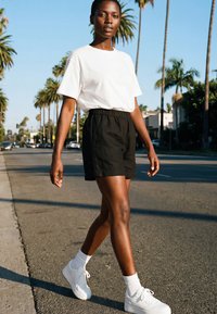 Jonge vrouw die op een zonnige straat met palmbomen loopt, gekleed in een wit T-shirt, zwarte shorts, witte sokken en witte sneakers.