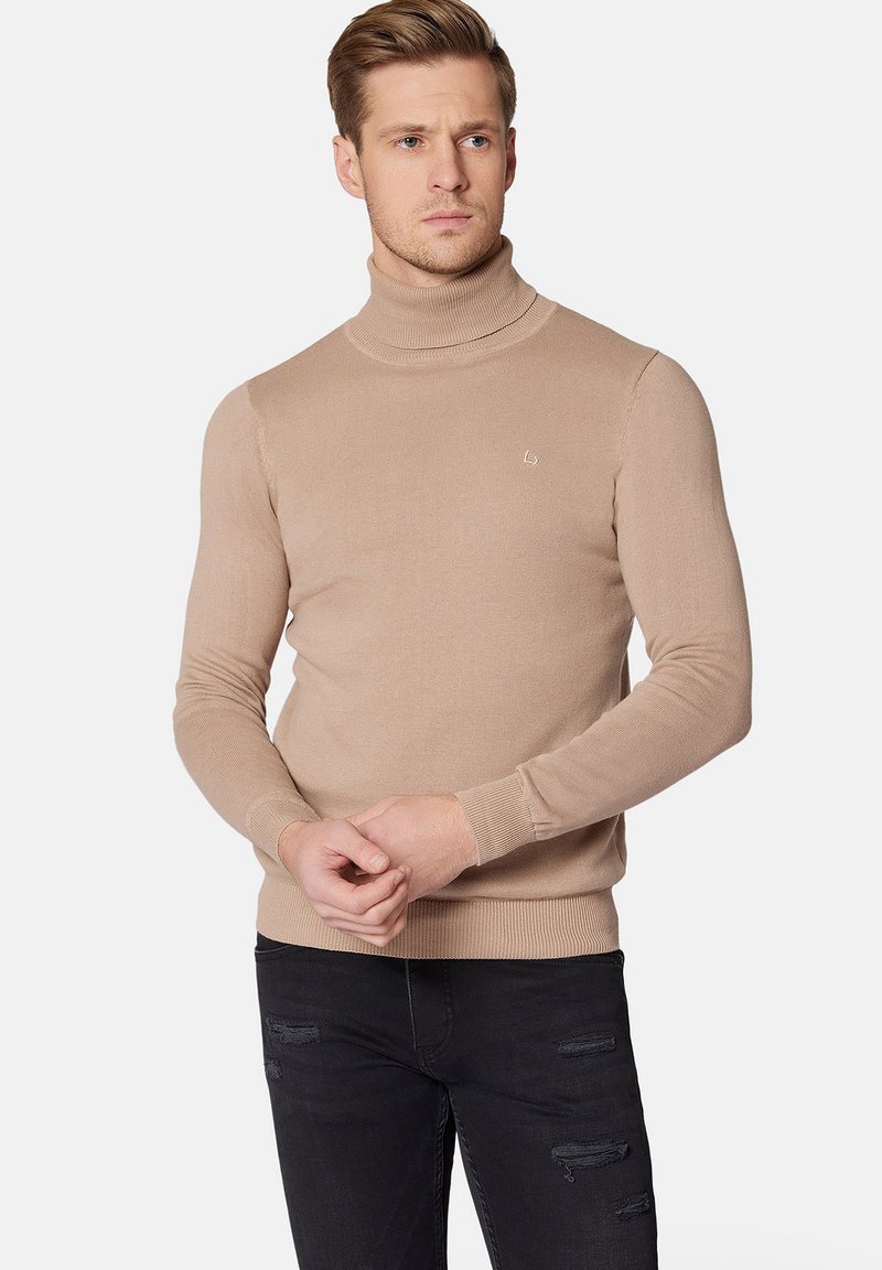 Beżowy sweter z golfem wykonany z dzianiny, o dopasowanym kroju i ściągaczach na rękawach, w połączeniu z czarnymi, postrzępionymi dżinsami.