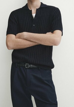 Homme les bras croisés, portant un polo en maille noire à manches courtes rentré dans un pantalon noir avec une ceinture noire sur un fond uni.