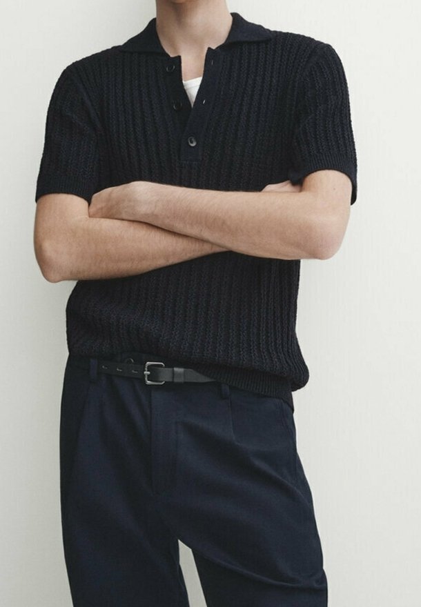 Homme les bras croisés, portant un polo en maille noire à manches courtes rentré dans un pantalon noir avec une ceinture noire sur un fond uni.