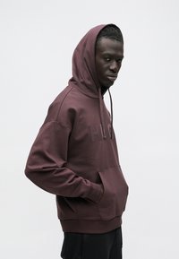 Felpa con cappuccio color bordeaux con tasca frontale, caratterizzata da un logo testurizzato sul petto. Vestibilità leggermente ampia con cappuccio regolabile e polsini a coste.