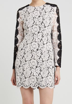 Witte bloemenkantjurk met zwarte kanten inzetten aan de lange mouwen, een geschulpte zoom en een ronde halslijn, gedragen door een staand persoon.