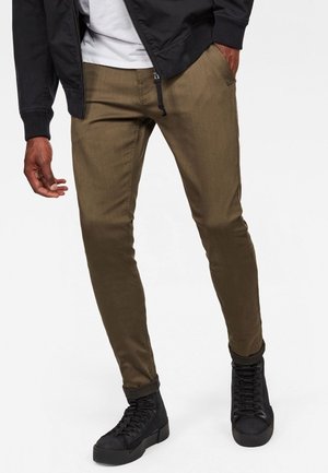Mann trägt schlanke olivgrüne Hose, schwarze hohe Sneakers, schwarze Jacke und weißes Hemd, steht mit einer Hand in der Tasche.