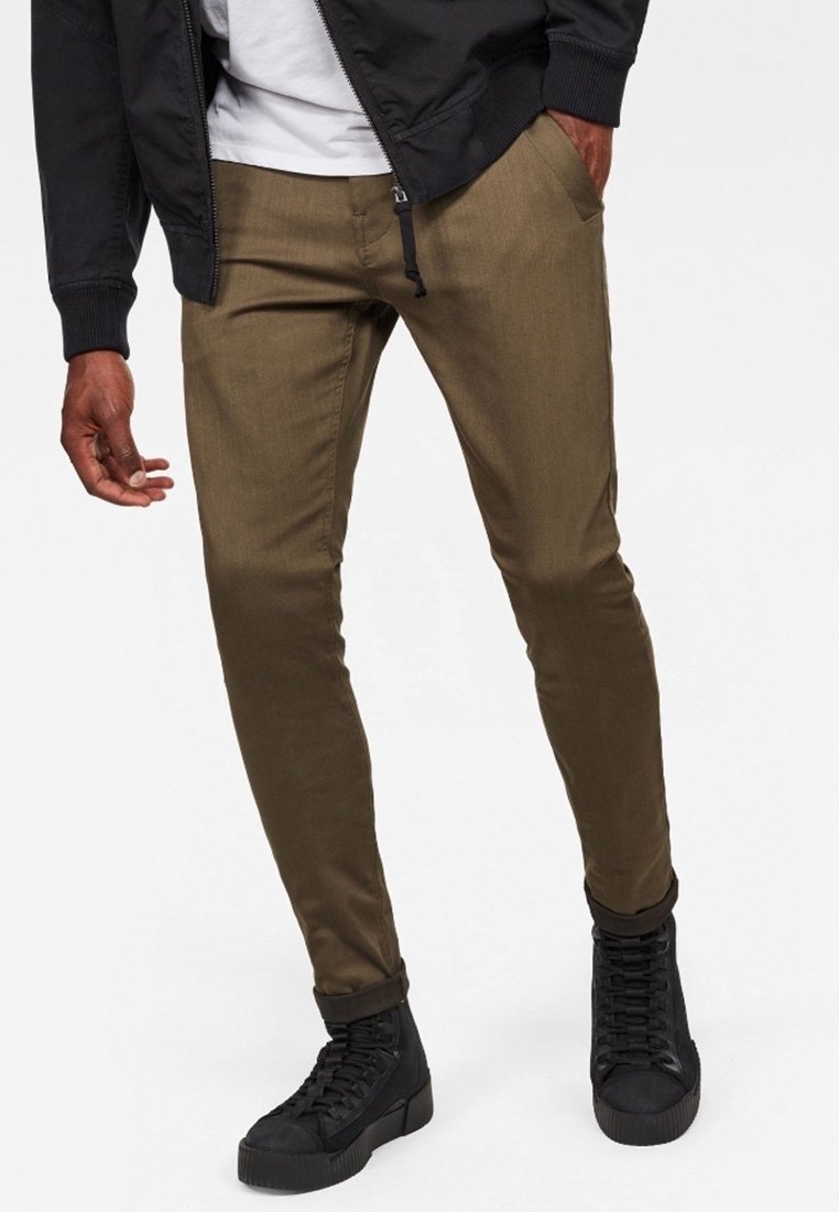 Homme portant un pantalon slim vert olive, des baskets montantes noires, une veste noire et une chemise blanche, debout avec une main dans la poche.