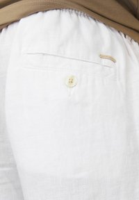 Un short en lin blanc avec une taille élastique, une poche arrière unique avec fermeture à bouton et une petite étiquette en tissu.