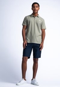 Polo shirt verde chiaro con dettagli in contrasto blu navy, abbinato a pantaloni corti blu navy. Il modello indossa scarpe da ginnastica bianche, in piedi contro uno sfondo chiaro.