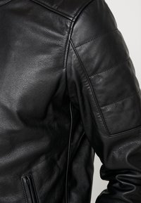 Giubbotto di pelle nera con materiale testurizzato, dettaglio trapuntato sulle spalle e tasche con zip per funzionalità. Design classico e aderente.