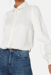 Blouse blanche à manches longues avec un devant boutonné, tissu doux et col classique. Associée à un jean en denim foncé mettant en valeur une silhouette ajustée.