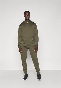 Mann steht in einem olivgrünen Hoodie und passenden Jogginghosen mit schwarzen Sneakers vor einem schlichten hellen Hintergrund.