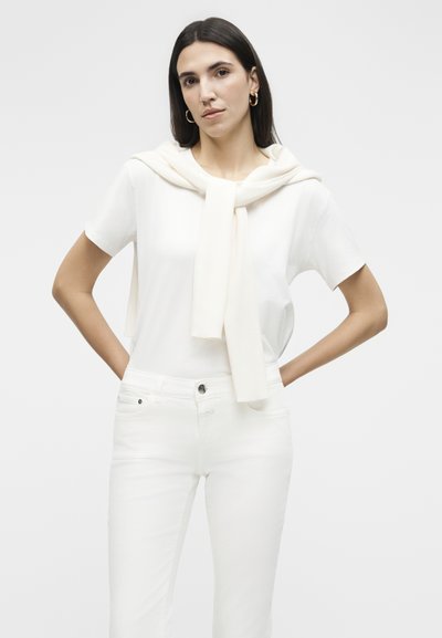 Femme aux cheveux raides et foncés portant un haut blanc à manches courtes, un jean blanc et un pull crème posé sur les épaules, les mains sur les hanches.