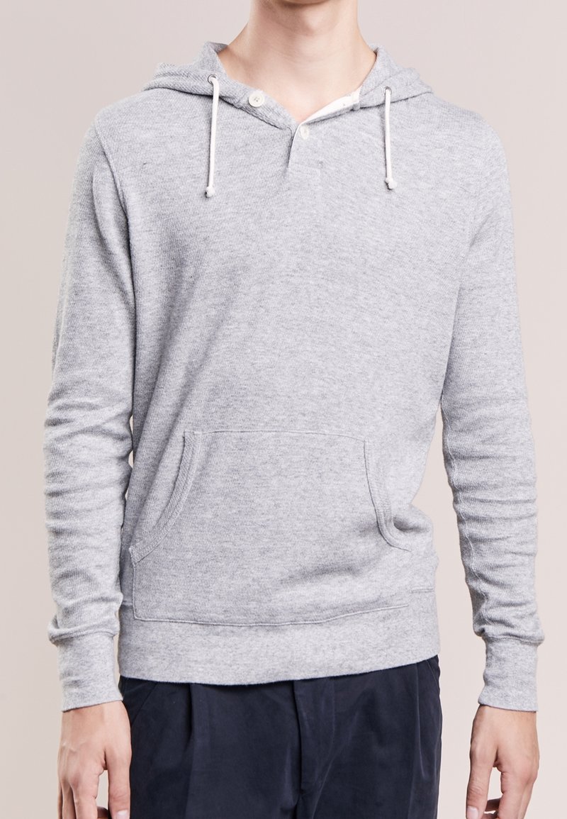 Personne portant un sweat à capuche gris clair avec poche avant, cordons blancs et pantalon noir sur un fond uni.
