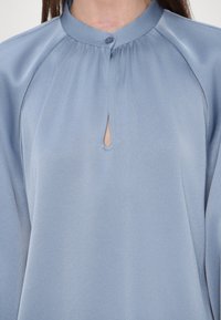 Femme portant un chemisier bleu clair à manches longues, une petite ouverture en forme de serrure à l'avant et un col rond avec une fermeture par un seul bouton.