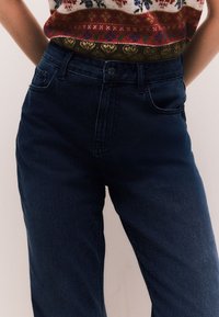 Tamsiai mėlynos spalvos džinsai iš džinso audinio, aukštu liemeniu, klasikiniu penkių kišenių dizainu ir subtilia tekstūra. Užsegami sagutėmis, juosmens diržo kilpos.