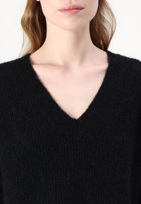 Pulover negru cu decolteu în V, confecționat dintr-un material tricotat texturat, având margini ribbate în jurul decolteului și un aspect moale, pufoș.