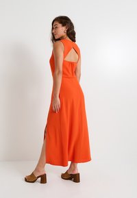 Vestido maxi de tela naranja brillante, con un diseño sin mangas, corte en la espalda y aberturas laterales. Combinado con sandalias de tacón marrones.