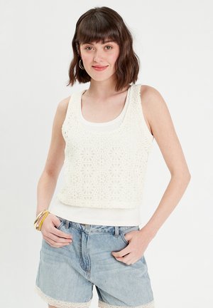 Witte gehaakte tanktop met een hexagonaal patroon en franjes onderaan, over een witte aansluitende blouse gedragen, gecombineerd met lichtblauwe denim shorts.