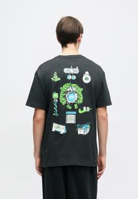 Μαύρο βαμβακερό t-shirt με πολύχρωμα καρτούν σχέδια στο πίσω μέρος, συμπεριλαμβανομένου ενός σχεδίου τοιχογραφίας και διάφορων παιχνιδιάρικων εικονιδίων.