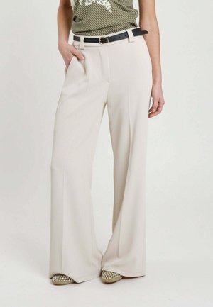 Donna che indossa pantaloni beige a gamba larga con vita alta, cintura nera e scarpe slip-on borchiate, mano in tasca, in piedi davanti a uno sfondo semplice.