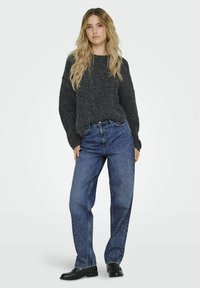 Pulover gri tricotat, asortat cu blugi albaștri cu accente cu cui. Ținuta este completată cu mocasini negri, pe un fundal simplu.