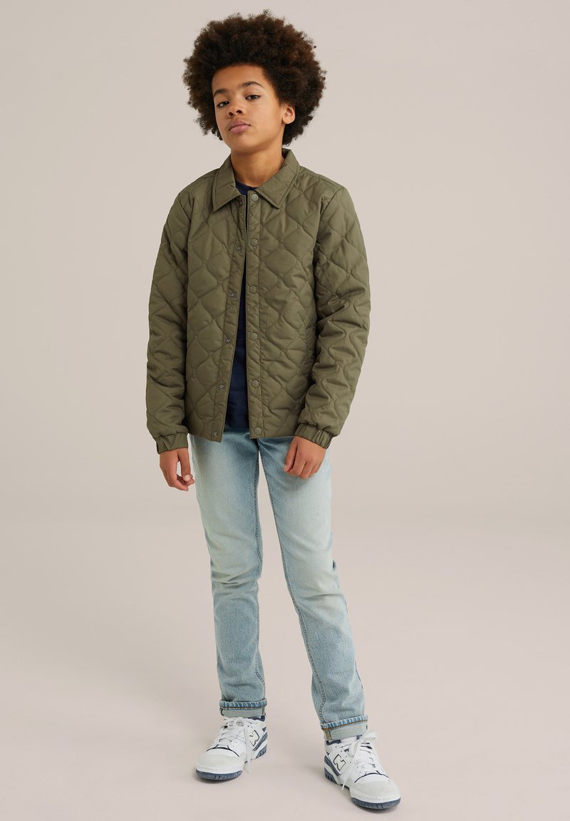 Quilted olive jacket with a collared neck and snap closures, paired with light blue jeans and white sneakers featuring blue accents.