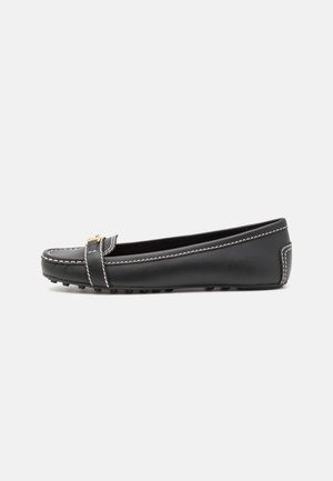 Sort læder flad loafer med hvide syninger og lille guldbeslag, gummisåle med nopper, set fra siden på ensfarvet baggrund.