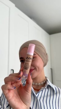Iemand met een beige hoofddoek en een gestreept shirt houdt de NYX Bare With Me concealer serum dicht bij de camera en glimlacht zachtjes.