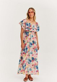 Kwiatowa sukienka maxi z falbaniem przy dekolcie bez ramion, ozdobiona różowymi różami i niebieskimi kwiatami na jasnym tle, połączona z brązowymi sandałami.