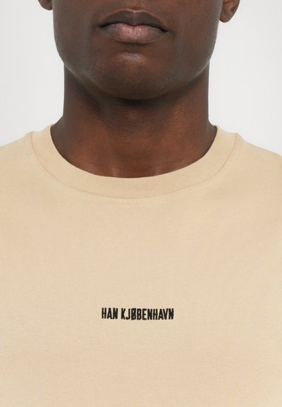T-shirt en coton beige avec col rond, affichant un petit texte noir "HAN KJØBENHAVN" centré sur la poitrine. Texture lisse, style ajusté.