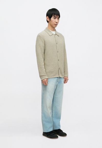 Béžový pletený sveter s golierom a zapínaním na gombíky, v kombinácii so svetlomodrými voľnými džínsami a čiernymi teniskami.