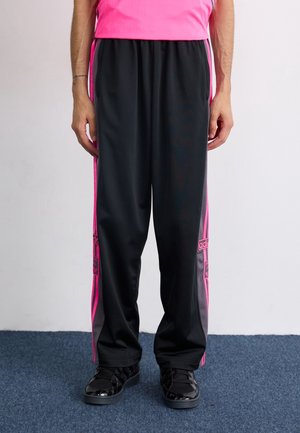 Personne portant un pantalon de survêtement Adidas noir et rose avec des chaussures, debout sur un sol recouvert d'un tapis bleu contre un mur blanc.