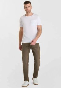 Weißes T-Shirt mit Rundhalsausschnitt und einer strukturierten Brusttasche, kombiniert mit khakifarbenen Hosen. Das Model trägt weiße Sneakers vor neutralem Hintergrund.