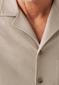 Camicetta beige con texture, colletto e bottoni, dotata di due bottoni e una tasca a toppa sul davanti.