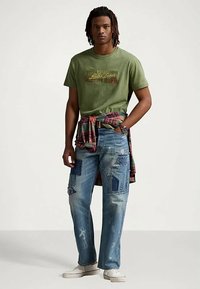 Πράσινο βαμβακερό μπλουζάκι με γραφικό σχέδιο, μπλε σκισμένα τζιν με πα patchwork, κόκκινο και πράσινο καρό πουκάμισο δεμένο στη μέση, λευκά παπούτσια.